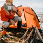 Mature man drinking hot drink in metal cup near tent on winter camping